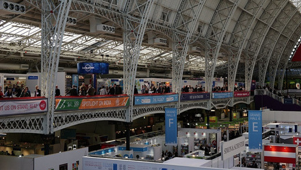 Wide-angle view of the London Book Fair inside a large exhibition hall with a glass and metal arched ceiling. The venue is bustling with attendees